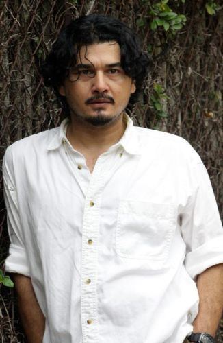 Azri Iskandar with a fearful look and both hands on his pocket while leaning on his right shoulder on a tree in the background, has black short hair, a beard, and mustache, and wearing a black watch and a white polo long sleeve