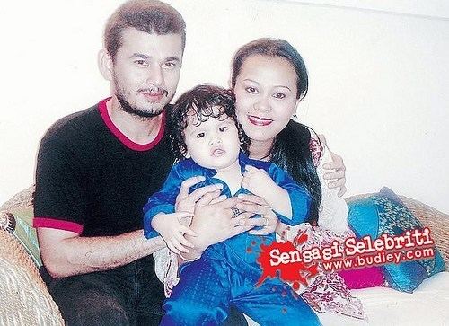 Azri Iskandar smiling while hugging her wife’s left shoulder, Ellie Suriaty and hugging their son in between and they are sitting on a couch with pillows on it. Azri has black hair, a beard, and a mustache wearing rings on both hand fingers, a bracelet, a black shirt and denim pants, and Ellie is smiling has black hair wearing a white printed long sleeve dress and their son is wearing a blue jumpsuit