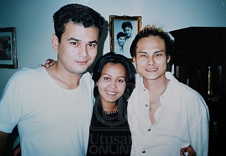 Azri Iskandar along with his wife, Ellie Suriaty, and a man are closed to each other with a picture frame in the background. Azri with a smirk on his face has black hair and wearing a silver necklace and a white shirt, Ellie is smiling, has black hair wearing a necklace and a black blouse, and the man beside Ellie is smiling and has black hair with blonde highlights and wears a silver necklace and white unbuttoned polo long sleeve