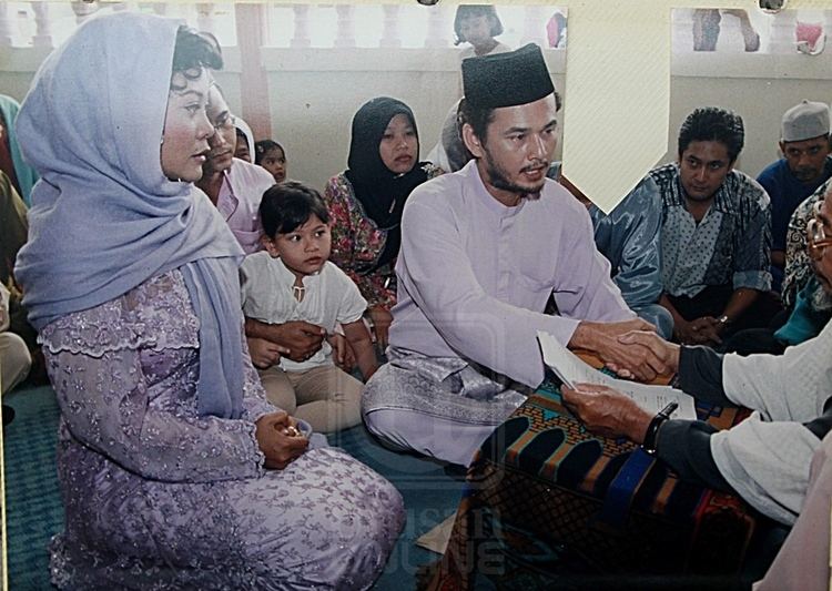 Azri Iskandar sitting and talking to someone while doing a handshake in his left hand to a man’s right hand with men and women and children around them in the background, the men are wearing kufi and polo long sleeves and women wears a hijab and a long sleeve dress. Azri has black hair and a beard and mustache, wearing a black kufi and a purple thobe