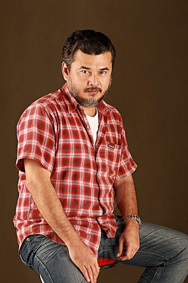Azri Iskandar with a fierce look and sitting on a red chair, has black hair, a beard and mustache wearing a watch on his left wrist, a white shirt under a red stripe polo, and denim pants