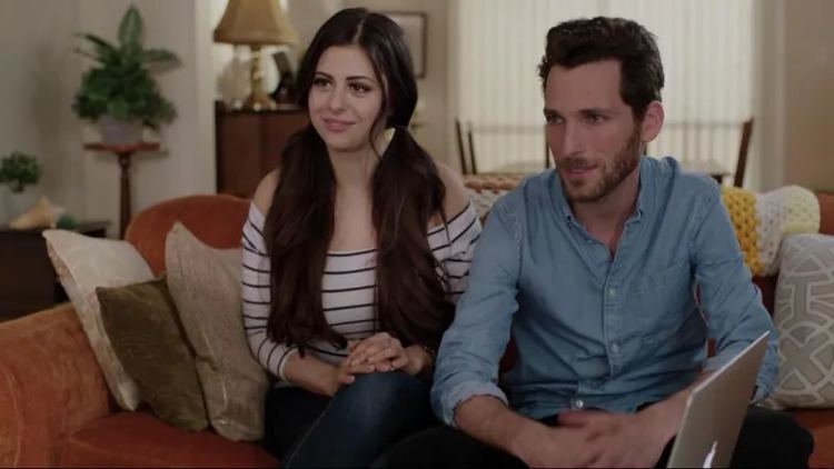 In a room with furniture, Azzyland on left is smiling, has long hair, both hands on her lap, sitting down on the orange couch with pillows on her right, colored white, brown, and light brown, she is wearing a white striped off-shoulder top and blue jeans. Ethan Cole on right is smiling, has black hair, a beard, and a mustache, sitting on an orange couch with a light brown, yellow, and white pillow with a design on his left and a MacBook in front, he is wearing blue long sleeves in a scene from My 90-Year-Old Roommate (TV Series  2016).