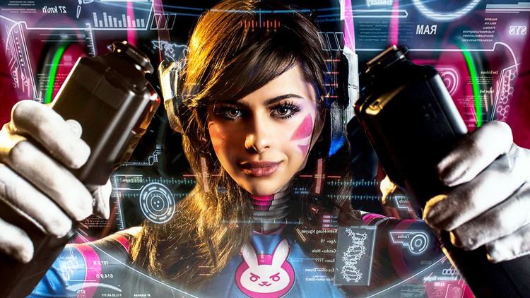 AzzyLand in D.Va Cosplay, with a serious face, wavy hair, a pink tattoo on her left cheek, with a monitor, while holding two black remotes, wearing headphones, white gloves, and a multi-colored cosplay costume with a bunny print in the center of D.Va.