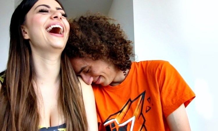 Azzyland on left is laughing has brown hair, wearing a cleavage showing top. On right is Jordi van den Bussche smiling and has brown curly hair, leaning on Azzyland, he wears an orange shirt with a print.