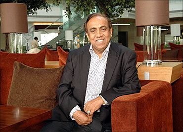 B. Ravi Pillai smiling while sitting on the couch and wearing a black coat and white long sleeves