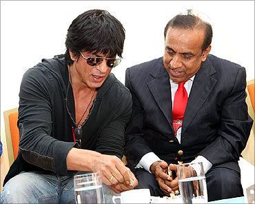 B. Ravi Pillai sitting on the chair with the man beside him while he is wearing a black coat, white long sleeves, and a red necktie