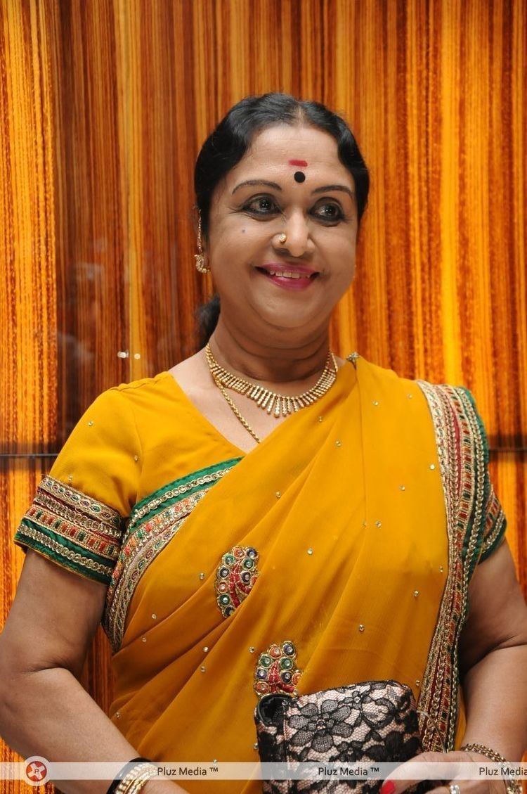 B. Saroja Devi smiling while wearing a yellow dress and some pieces of gold jewelry