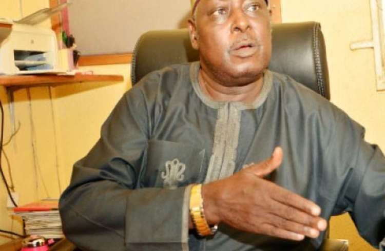 Babachir David Lawal the Secretary to the Government of the Federation is serious, sitting down on a black chair, has a printer and a pile of paper behind on his right, right palm up, wearing a gold wristwatch on his right hand, a gray dashiki agbada with a minimal design on his right pocket.