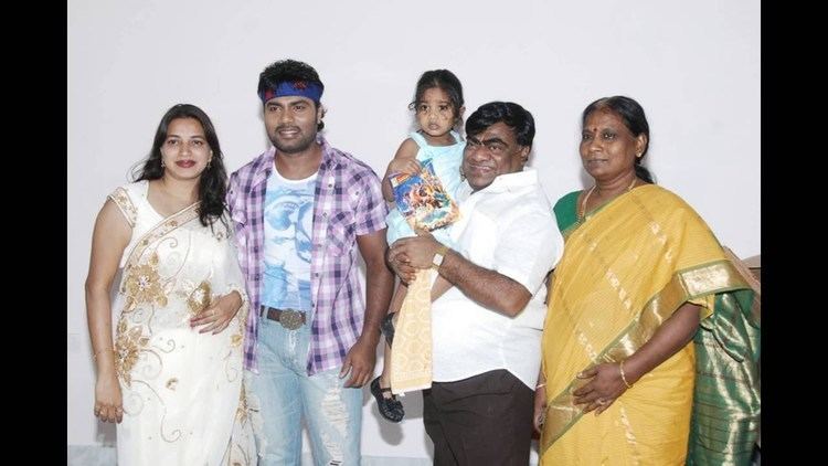 Babu Mohan smiling and carrying a toddler together with his family while he is wearing a white polo and black pants