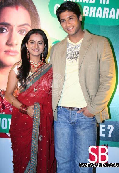 Aastha Chaudhary smiling and wearing a green and red dress while Sidharth Shukla wearing a beige coat, yellow t-shirt, and denim pants