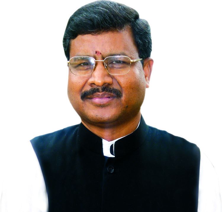 Babulal Marandi, Former Chief Minister of Jharkhand is smiling, and has black hair, a tika on his forehead, a mustache, and a mole on his left lower cheek, he wears eyeglasses and a black suit with a white collar suit.