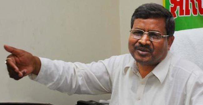 Babulal Marandi, Former Chief Minister of Jharkhand is serious, has black hair, and a mustache, right hand up, and behind him is a poster with red, green, and yellow prints, he is wearing a ring on his right hand, eyeglasses, and white long sleeves.