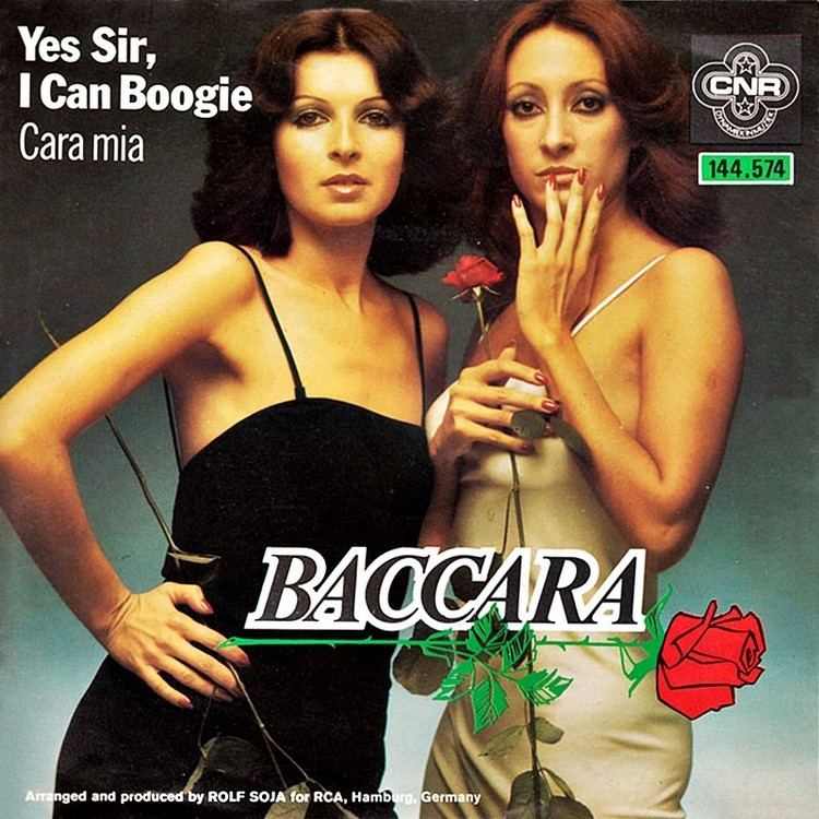 Mayte Mateos and María Mendiola of the Baccara vocal duo are holding a rose while Mayte is wearing a black sleeveless dress and María is wearing a cream sleeveless dress