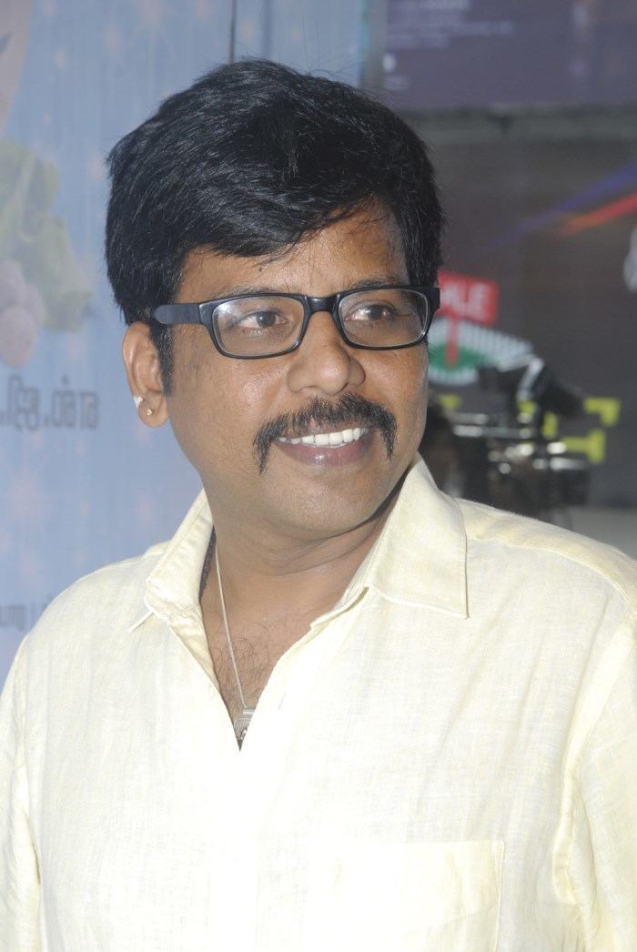 Badava Gopi smiles while wearing a collared shirt, a necklace, and a pair of eyeglasses