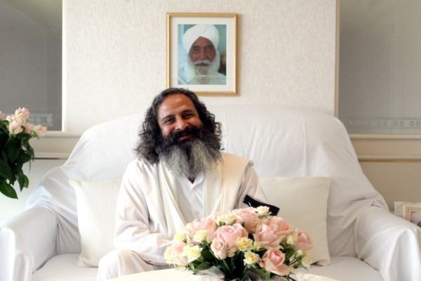 Baljit Singh (Sant Mat) sitting on a couch wearing an all-white shirt while holding a flower bouquet | with a picture frame of Sant Thankar Singh at his back