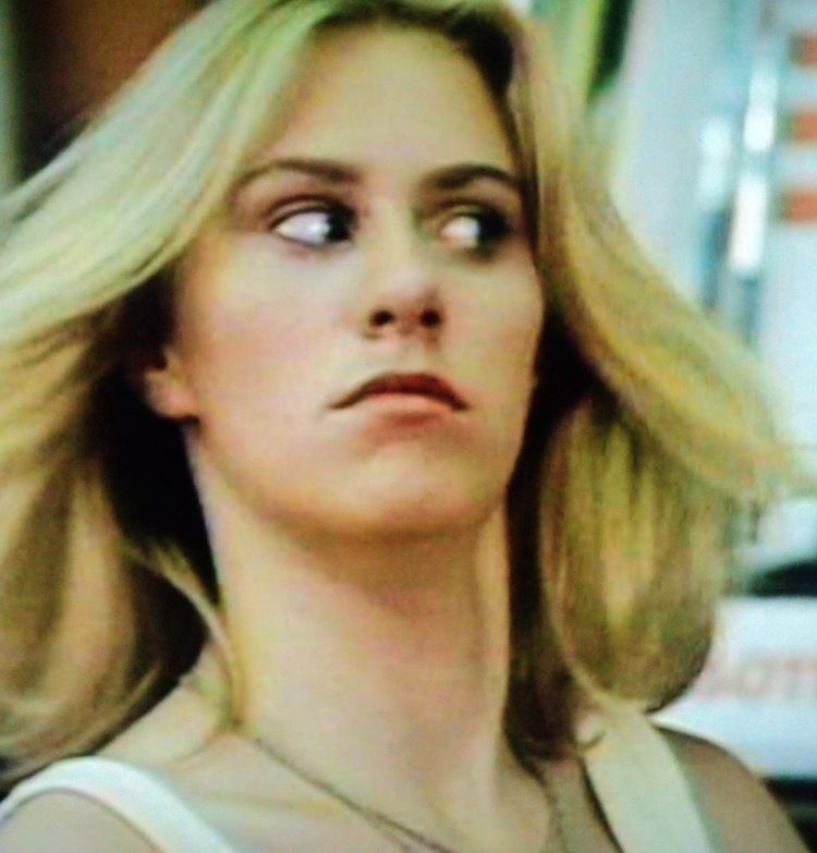 Bambi Woods with blonde hair and a weird face, wearing a white sleeveless top.