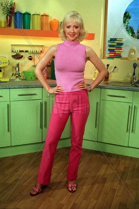 Barbara Durkin smiling while standing and holding her hips with both hands inside of a kitchen and kitchen tools in the background, in a TV show “My Parents Are Aliens cast: where are they now?”,  she has blonde hair, wearing an earrings, a watch on her left hand, pair of sandals, a pink sleeveles neck top and pink pants