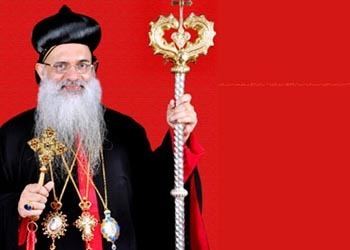 Baselios Mar Thoma Paulose II smiling while holding a blessing cross and wearing a black and red cassock and necklaces