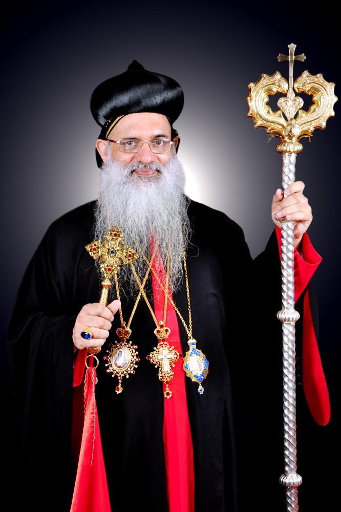 Baselios Mar Thoma Paulose II smiling while holding a blessing cross and wearing a black and red cassock and necklaces