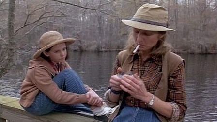 Jena Malone as Ruth Anne sitting and holding her hand on her leg while looking at Glenne Headly as Ruth who is lighting her cigarette with a stainless lighter with bare trees and a lake in the background, in a scene from the 1996 American drama film, Bastard Out of Carolina. Jena is wearing a beige hat, white socks, a pair of black sneakers, and blue denim pants. While Glenne is wearing a brown hat, a silver wristwatch on her left, a black bracelet on her right wrist, wearing a pair of blue denim pants, and a brown vest over a brown checkered collared long sleeve.