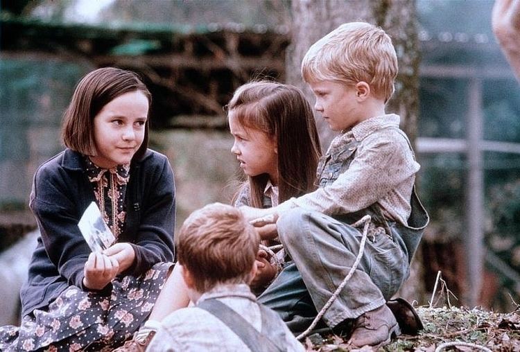 Jena Malone as Ruth, Lindley Mayer as Reese, Jamison Stewart as Grey, and Timothy Stewart as Garvey (from left to right) sitting while looking at each other and talking in a scene from the 1996 American drama film, Bastard Out of Carolina. Jena has short brown hair tucked in her ears, wearing a blue dress with prints under a black collared blazer; Lindley has long and straight brown hair clipped on the side, wearing a collared dress; Jamison has blonde hair, wearing a pair of old leather shoes, and a dirty collared sleeve shirt under a gray denim jumper; and Timothy in brown hair is wearing a grayish collared shirt inside his dark gray denim jumper.