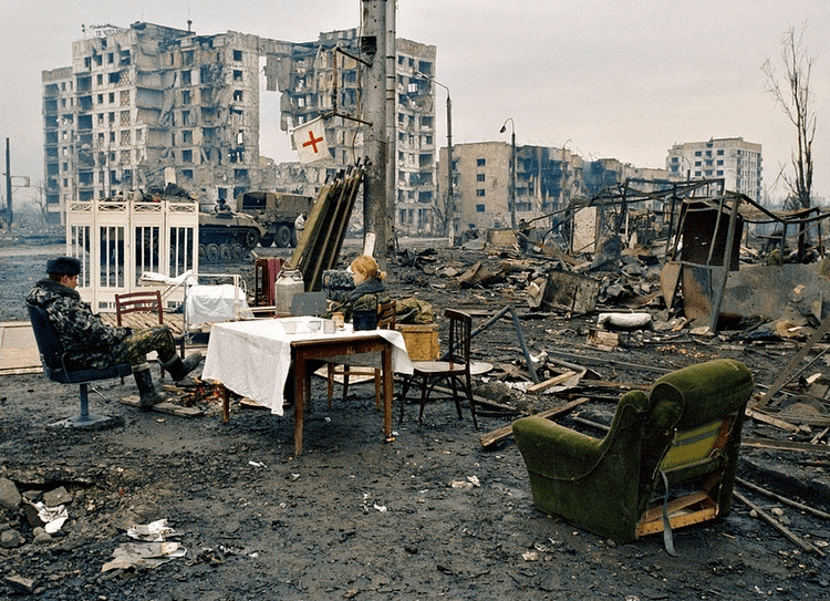 Russian soldiers sitting on the chair and resting at Minutka square, in Grozny, Chechnya with crippled & burnt buildings in the background while they are wearing camouflage trousers and jackets.