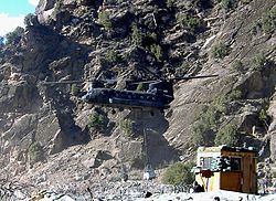 A Chinook helicopter lands at Combat Outpost Keating, Afghanistan, has a rock a wall with barbed wires and a small yellow barracks.