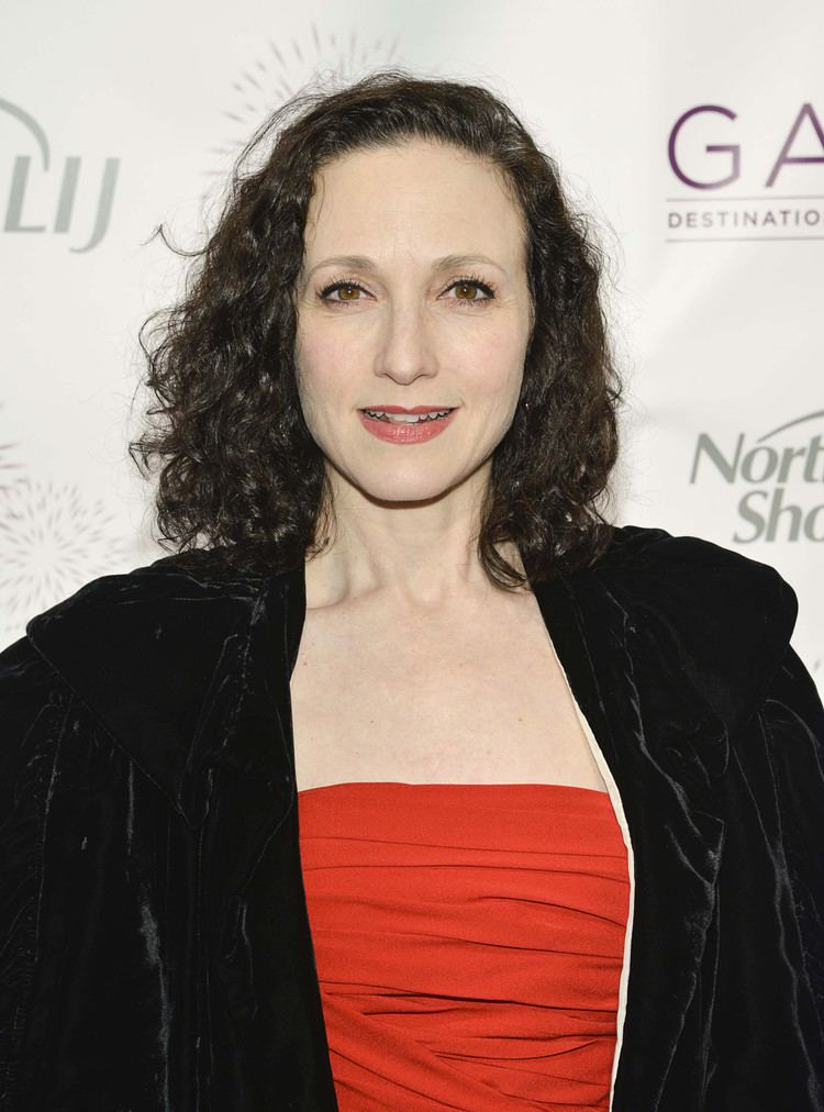 Bebe Neuwirth smiling, with curly hair and wearing a black jacket and a red top.