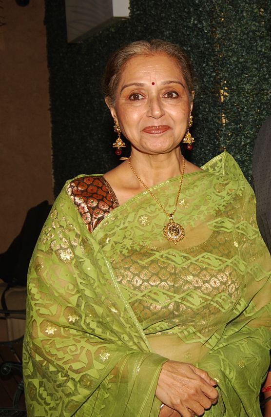 Beena Banerjee smiling while wearing a brown dress, green shawl, gold necklace, and earrings