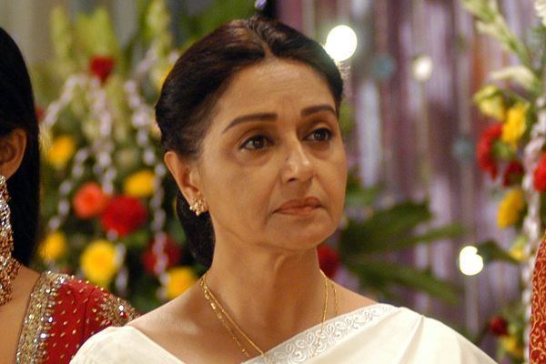 Beena Banerjee looking afar while wearing a white dress, gold necklace, and earrings