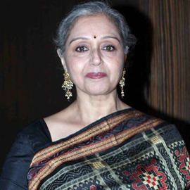 Beena Banerjee with a tight-lipped smile while wearing a black dress, colorful dupatta, and gold earrings