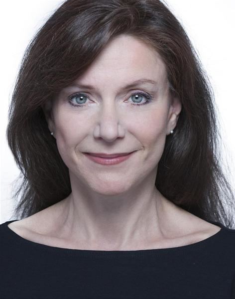 Belinda Lang with a tight-lipped smile, wavy hair, wearing earrings and a black top.