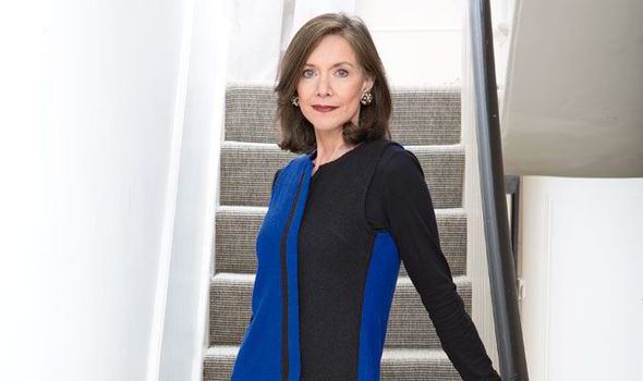 Belinda Lang with a serious face while standing on a stair, with shoulder-length hair, wearing earrings, and a black and blue long sleeve dress.