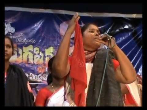 In the middle Belli Lalitha, an Indian Folk, is serious, singing, right hand up with a scarf on her pinky, while left hand holding the mic, has black hair, wearing black and red indian dress, around her is supporters with blue tarpaulin in the back.