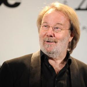 Benny Andersson smiling while wearing a black coat, black long sleeves, and eyeglasses
