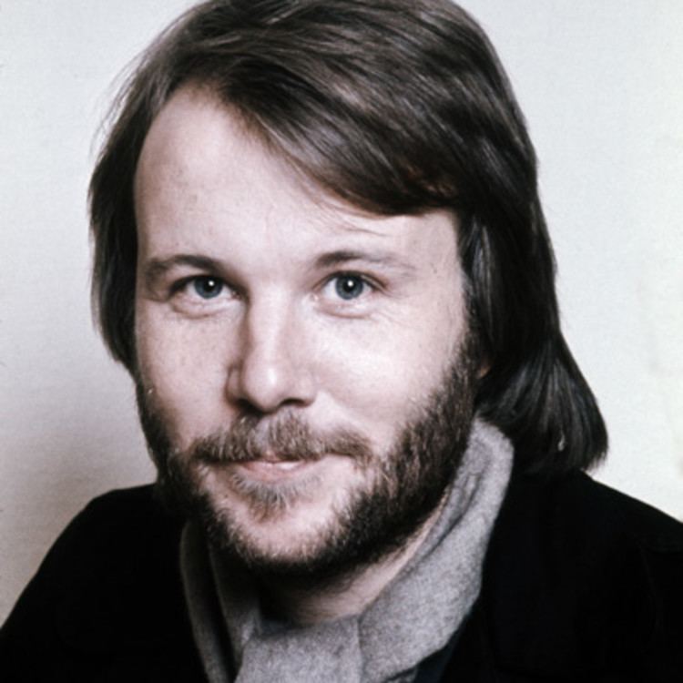 Benny Andersson smiling with mustache and beard while wearing a black shirt and gray scarf