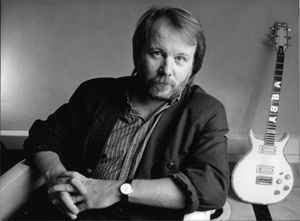 Benny Andersson sitting on the chair beside a guitar while wearing a black coat and striped long sleeve