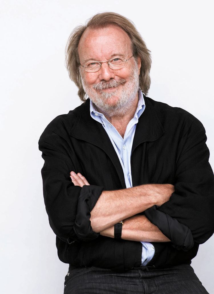 Benny Andersson smiling while arms crossed and wearing eyeglasses, a black coat, and blue long sleeve