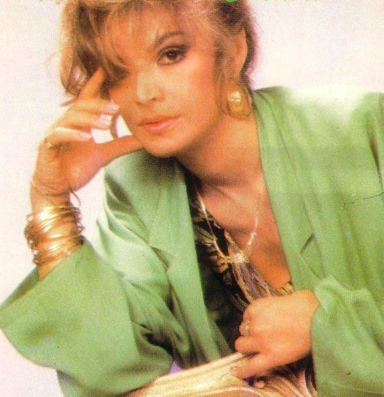 Bergen looking fierce with blonde hair covering her right eye and wearing a colored blouse under a green coat, gold necklace, gold earrings, gold bracelets, and finger rings