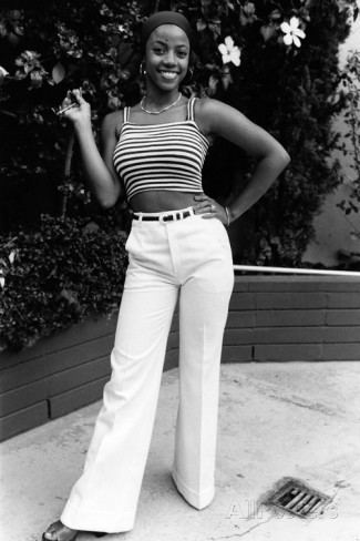 Bern Nadette Stanis smiling while hand on her hips and wearing a striped sleeveless top, pants, necklace, and bracelet