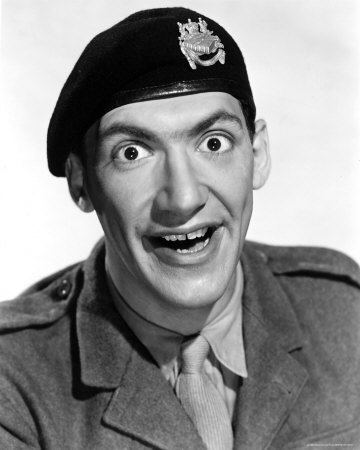 Bernard Bresslaw with a funny face, wearing a black flat cap with a badge, a gray coat over gray long sleeves, and a gray necktie.