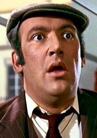 Bernard Bresslaw with a shocked face, wearing a gray flat cap, red coat over white long sleeves, and red necktie.