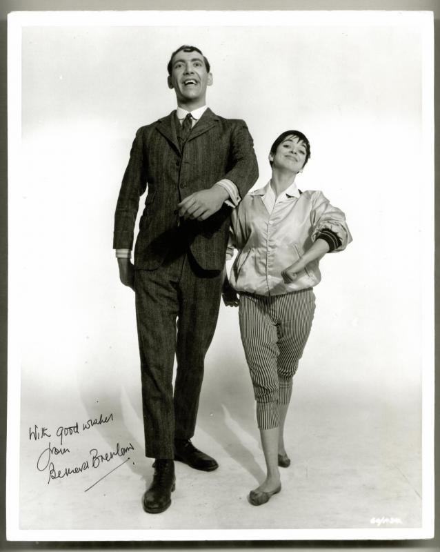 Bernard Bresslaw is smiling while swinging her arms with a girl on his side with an autograph on the lower-left corner. Bernard is wearing a black coat over white long sleeves, a black necktie, black pants, and black shoes while the girl is wearing a silk jacket, striped pants, and black shoes.