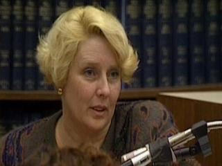 Betty Broderick with blonde hair, a microphone in front of her, and lot of books behind her.