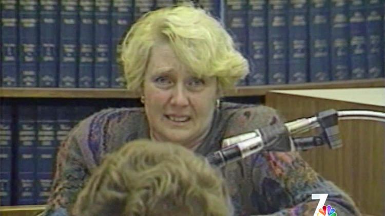 Betty Broderick with blonde hair, a microphone in front of her, and lot of books behind her.