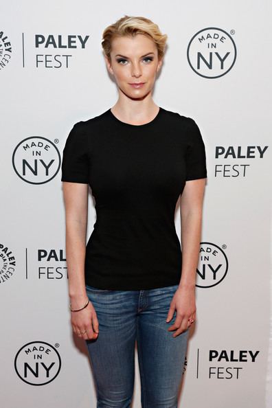 Betty Gilpin with a tight-lipped smile while wearing a black blouse and denim pants