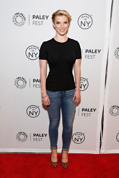 Betty Gilpin smiling while wearing a black blouse and denim pants