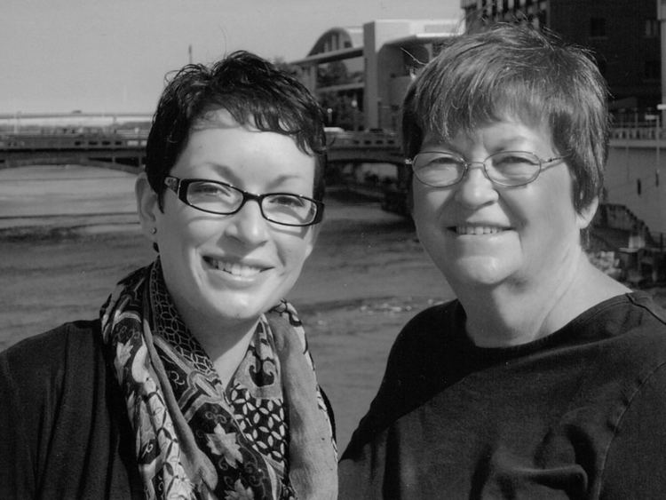 Mahtob and Betty Mahmoody are smiling. Mahtob with short hair, wearing eyeglasses, a scarf, and a black shirt while Betty wearing eyeglasses and a black shirt.