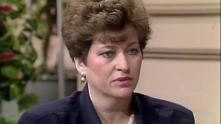 Betty Mahmoody with a serious face, short hair, wearing pearl earrings, a necklace, and a blue blouse.