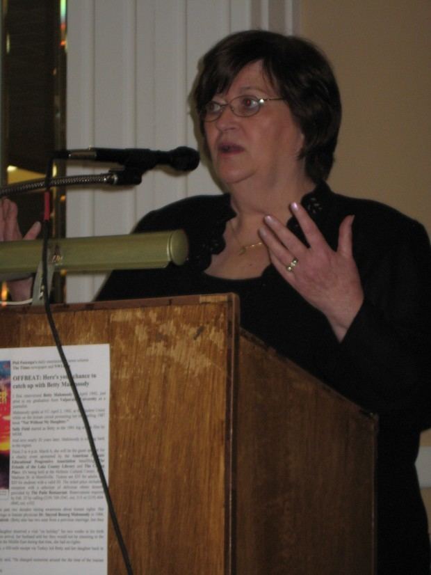 Betty Mahmoody with a serious face in front of a podium and microphones, wearing eyeglasses, and a black long sleeve top.